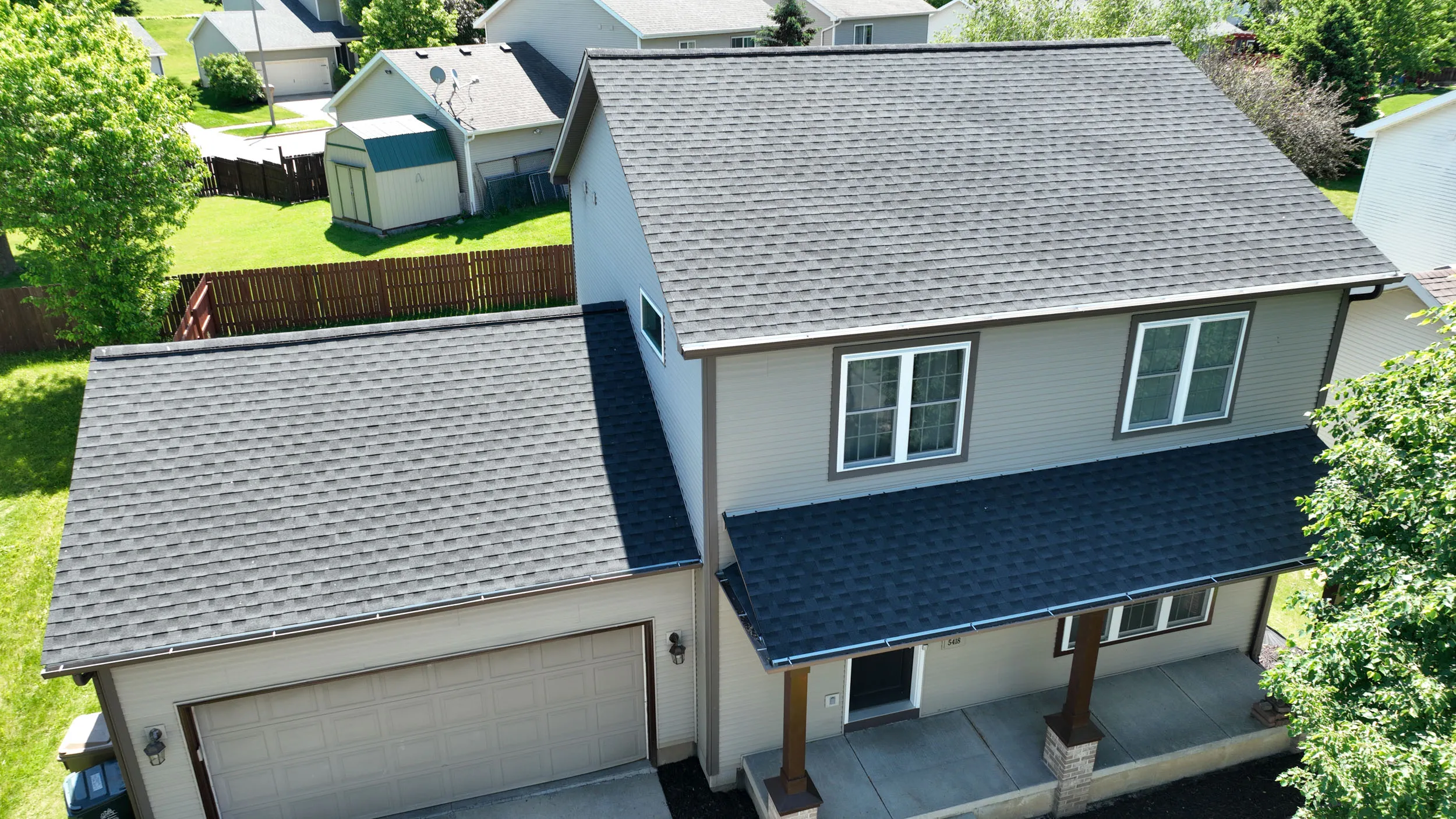 A two-story house with beige siding, gray shingle roofs, and an attached garage, surrounded by trees and a fenced lawn.
